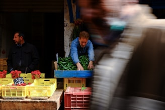 A bustling market scene with two men near a vegetable stall. One man is arranging green peppers in a blue crate, while the other stands beside piles of strawberries in yellow containers. A blur of motion suggests people walking by.