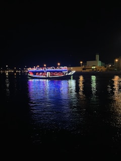 A boat adorned with bright LED lights in various colors is sailing on a calm body of water at night. The lights reflect vibrantly on the surface of the water, creating a mesmerizing pattern. In the background, there is a shoreline with buildings and streetlights illuminating the scene.