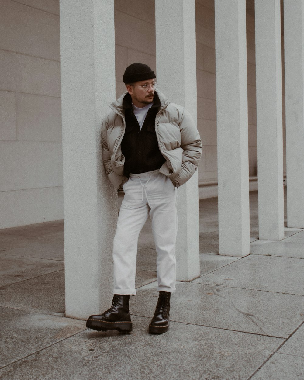 Hombre con blazer gris y pantalones blancos de pie junto a un poste de concreto blanco durante el día
