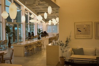 An aesthetically pleasing cafe featuring large, modern, white pendant lights hanging from the ceiling. Long communal tables with wooden chairs are lined up parallel to a series of large windows on one side. A cozy seating area is on the right with a sofa and decorative plants, complemented by minimalistic art on the walls.