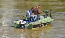 A small fishing boat with three people onboard is on a calm body of water. The boat is equipped with fishing gear and has decals on its side. The water appears slightly brownish, indicating a river or lake setting.