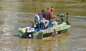 A small fishing boat with three people onboard is on a calm body of water. The boat is equipped with fishing gear and has decals on its side. The water appears slightly brownish, indicating a river or lake setting.