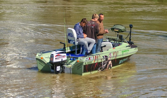 A small fishing boat with three people onboard is on a calm body of water. The boat is equipped with fishing gear and has decals on its side. The water appears slightly brownish, indicating a river or lake setting.