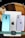 Several electronic devices including a tablet and two smartphones are arranged on a surface. The tablet has an Apple logo, indicating it is likely an iPad. One smartphone is a OnePlus with a blue back, and the other is an Apple iPhone with a light purple back. At the bottom, there is a smartwatch with a square face on a green band.