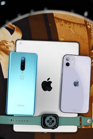 Several electronic devices including a tablet and two smartphones are arranged on a surface. The tablet has an Apple logo, indicating it is likely an iPad. One smartphone is a OnePlus with a blue back, and the other is an Apple iPhone with a light purple back. At the bottom, there is a smartwatch with a square face on a green band.