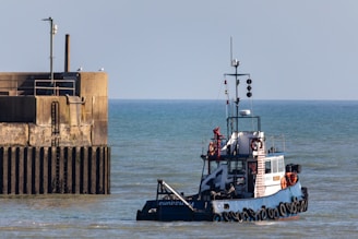 A small tugboat navigates the calm waters near a concrete pier structure, where a couple of seagulls perch. The sky is clear, and there are navigational and communication equipment visible on the boat.