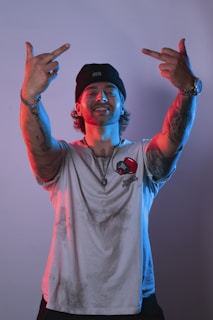 A person wearing a beanie and a T-shirt with tattoos on their arms, showing a confident expression. The lighting gives a dramatic effect with blue and red tints.