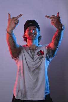 A person wearing a beanie and a T-shirt with tattoos on their arms, showing a confident expression. The lighting gives a dramatic effect with blue and red tints.