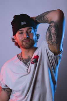 A person wearing a beanie and a white t-shirt with a red design on it is posing with one arm raised, revealing a detailed tattoo on the inner arm. The lighting includes shades of blue and pink, creating a modern, edgy atmosphere.