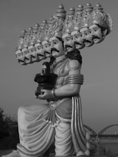 A large statue of a mythological figure with ten heads and intricately detailed headgear, holding a cylindrical object. The statue features elaborate drapery and is set against a backdrop of a bridge and trees.