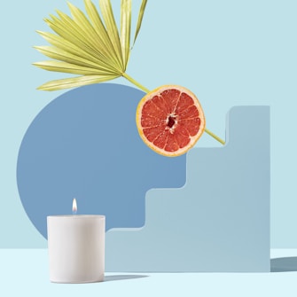 A minimalistic arrangement with a white candle, a sliced grapefruit, and a green palm leaf placed on a geometric blue structure against a light blue background.