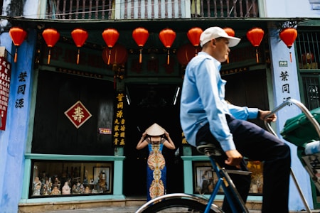 A shop with traditional architecture is adorned with bright red lanterns. In front of the entrance, a woman wearing a colorful dress and a conical hat stands with her back to the viewer. A man on a bicycle, dressed in a light blue shirt and white cap, moves across the foreground. The shop windows display various figurines and decorations, while signs with vertical text are visible.