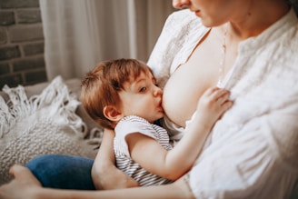 A close-up image of a mother breastfeeding her baby. The child is wearing a striped shirt, and the scene conveys a sense of intimacy and nurturing. Soft lighting and neutral tones create a warm atmosphere.