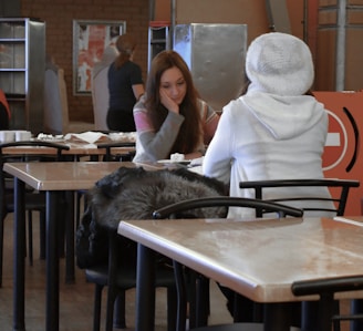 Two women are seated at a table in a casual dining setting. One woman faces the camera with her hand resting on her chin, appearing thoughtful. The other woman sits with her back to the camera, wearing a white sweater and hat. The table has remnants of food and drinks, and the atmosphere suggests a relaxed, informal conversation. In the background, other patrons and furniture are visible.