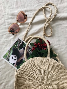 A woven straw handbag is placed on a beige surface. It contains green leaves and red berries peeking out from the top. Next to the bag, there is a pair of sunglasses with rose-tinted lenses reflecting a person taking a photo. A photograph of a panda climbing a tree trunk is partially tucked under the bag.