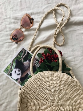 A woven straw handbag is placed on a beige surface. It contains green leaves and red berries peeking out from the top. Next to the bag, there is a pair of sunglasses with rose-tinted lenses reflecting a person taking a photo. A photograph of a panda climbing a tree trunk is partially tucked under the bag.