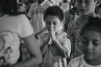 Children stand with their eyes closed, appearing to be praying or deep in thought. One child in the center is holding their hands together in a prayer gesture. The background is out of focus, with other children also visible.