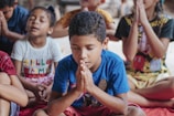 A group of children sitting cross-legged with their eyes closed and hands pressed together in a prayer-like position. The children are wearing colorful clothes and appear to be in a calm and focused state.