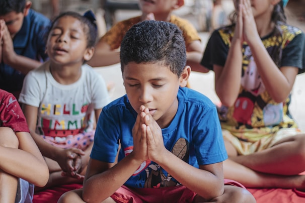 A group of children sitting cross-legged with their eyes closed and hands pressed together in a prayer-like position. The children are wearing colorful clothes and appear to be in a calm and focused state.