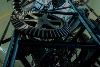 A complex arrangement of large, interlocking gears and mechanical components, predominantly made of metal, viewed from a low angle. The gears have thick teeth and are set against a backdrop of a bright, dome-like ceiling, creating a contrast between the dark machinery and the light background.