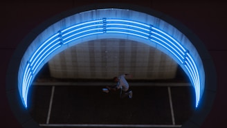 Aerial view of a person performing a dynamic jump or dance move beneath a circular structure illuminated by bright blue neon lights. The scene appears to be set in an urban environment, potentially within a tunnel or underpass with modern design elements.