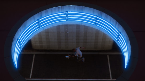 Aerial view of a person performing a dynamic jump or dance move beneath a circular structure illuminated by bright blue neon lights. The scene appears to be set in an urban environment, potentially within a tunnel or underpass with modern design elements.