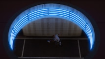 Aerial view of a person performing a dynamic jump or dance move beneath a circular structure illuminated by bright blue neon lights. The scene appears to be set in an urban environment, potentially within a tunnel or underpass with modern design elements.