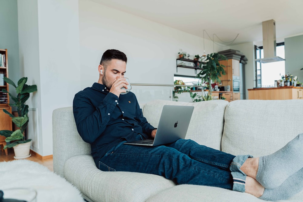 man in blue jacket and blue denim jeans sitting on gray sofa using macbook