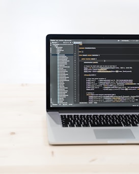 A laptop is placed on a light wooden table, displaying a code editor with PHP code. The screen shows the directory structure on the left and code written in the main editor area on the right.