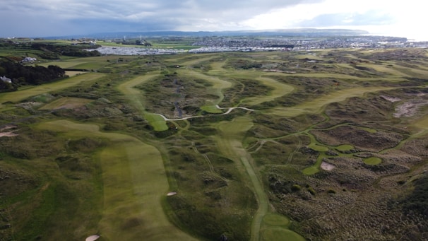 An expansive landscape view of a golf course featuring undulating greens, sand bunkers, and pathways. The course is situated in a coastal area with a distant view of the sea and a small town. Hilly terrain surrounds the course, and the sky is partly overcast with patches of sunlight illuminating portions of the grass.