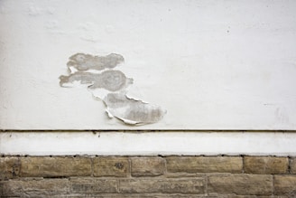 A wall with a section of peeling white paint, exposing the rough surface beneath. The wall consists of a smooth, white-painted upper area and a lower section with textured, beige stone-like bricks. The peeling paint reveals a greyish underlayer and adds a sense of deterioration.