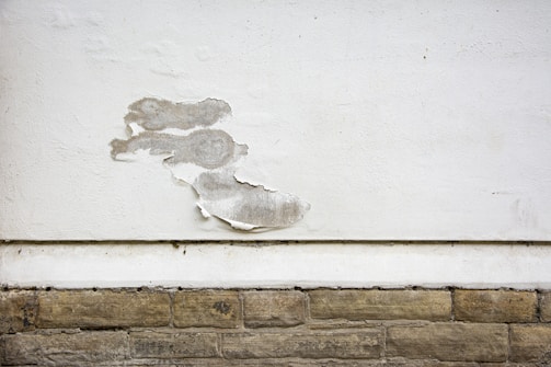 A wall with a section of peeling white paint, exposing the rough surface beneath. The wall consists of a smooth, white-painted upper area and a lower section with textured, beige stone-like bricks. The peeling paint reveals a greyish underlayer and adds a sense of deterioration.