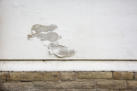 A wall with a section of peeling white paint, exposing the rough surface beneath. The wall consists of a smooth, white-painted upper area and a lower section with textured, beige stone-like bricks. The peeling paint reveals a greyish underlayer and adds a sense of deterioration.