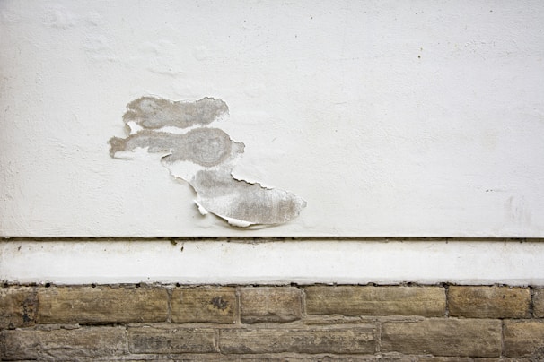 A wall with a section of peeling white paint, exposing the rough surface beneath. The wall consists of a smooth, white-painted upper area and a lower section with textured, beige stone-like bricks. The peeling paint reveals a greyish underlayer and adds a sense of deterioration.
