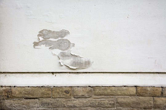 A wall with a section of peeling white paint, exposing the rough surface beneath. The wall consists of a smooth, white-painted upper area and a lower section with textured, beige stone-like bricks. The peeling paint reveals a greyish underlayer and adds a sense of deterioration.