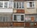 A worn-out facade of a building with a variety of mismatched windows and materials. The surfaces show signs of rust and weathering, with some windows featuring metal grilles and others partially boarded up. A 'for sale' sign with contact information is visible on one of the sections.