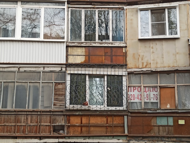 A worn-out facade of a building with a variety of mismatched windows and materials. The surfaces show signs of rust and weathering, with some windows featuring metal grilles and others partially boarded up. A 'for sale' sign with contact information is visible on one of the sections.