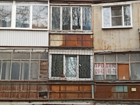 A worn-out facade of a building with a variety of mismatched windows and materials. The surfaces show signs of rust and weathering, with some windows featuring metal grilles and others partially boarded up. A 'for sale' sign with contact information is visible on one of the sections.