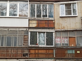 A worn-out facade of a building with a variety of mismatched windows and materials. The surfaces show signs of rust and weathering, with some windows featuring metal grilles and others partially boarded up. A 'for sale' sign with contact information is visible on one of the sections.