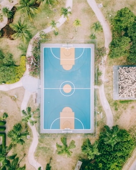 An aerial view of an outdoor basketball court situated in a park-like area. The court has a blue playing surface with tan areas for the key and three-point arcs. Surrounding the court are pathways, lush green trees, and some shrubbery. There is also a small building with a tiled roof to the right side of the image.