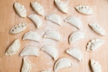 Multiple uncooked empanadas are arranged on a floured wooden surface. Each empanada is folded and sealed in a variety of styles, with some having intricate braided edges and others simple, rounded edges. The background is lightly dusted with flour, adding to the preparation theme.