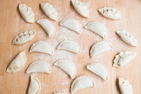 Multiple uncooked empanadas are arranged on a floured wooden surface. Each empanada is folded and sealed in a variety of styles, with some having intricate braided edges and others simple, rounded edges. The background is lightly dusted with flour, adding to the preparation theme.