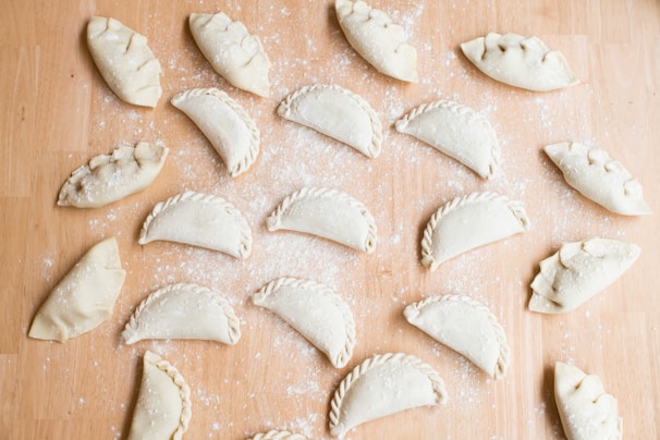 Multiple uncooked empanadas are arranged on a floured wooden surface. Each empanada is folded and sealed in a variety of styles, with some having intricate braided edges and others simple, rounded edges. The background is lightly dusted with flour, adding to the preparation theme.