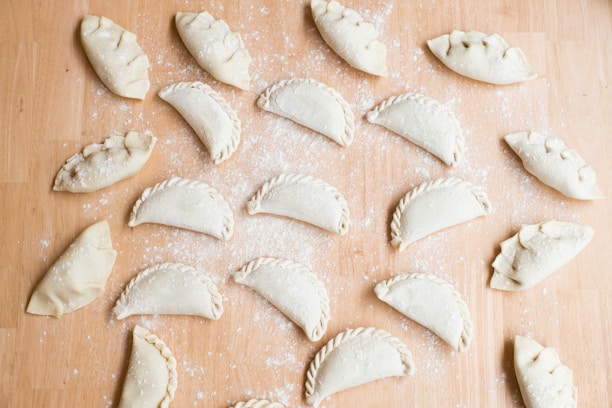 Multiple uncooked empanadas are arranged on a floured wooden surface. Each empanada is folded and sealed in a variety of styles, with some having intricate braided edges and others simple, rounded edges. The background is lightly dusted with flour, adding to the preparation theme.