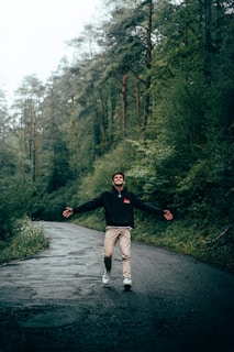 A person is joyfully walking along a wet, winding road surrounded by lush green trees in a forest setting. The mood is one of freedom and happiness.