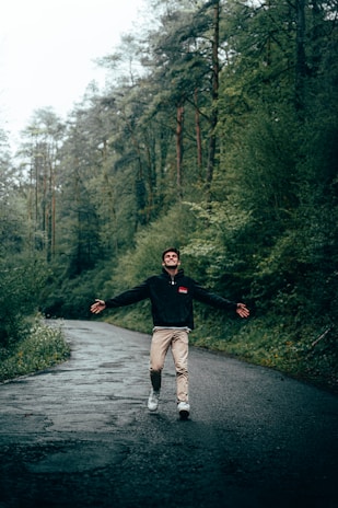 A person is joyfully walking along a wet, winding road surrounded by lush green trees in a forest setting. The mood is one of freedom and happiness.