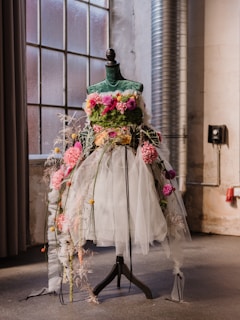 A dress form adorned with vibrant flowers and greenery, featuring a bodice covered in lush green foliage and pink blooms. The skirt is made of layered tulle and adorned with multiple colorful flowers, giving it a whimsical and artistic appearance. The setting is an industrial-styled room with large windows and a visible metallic vent, which contrasts with the delicate nature of the floral dress.