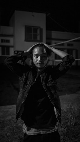 A person stands outdoor at night with eyes closed and hands placed on their head, appearing deep in thought or contemplation. The scene is primarily lit in grayscale, allowing for a strong focus on the person's expression and posture. The background shows a residential building that is dimly lit.