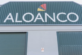 A large industrial building facade with the word 'ALOANCO' prominently displayed in white on a green background. Above the word, a multi-colored logo features a red, yellow, and orange triangular design. The building has green corrugated metal doors, and a small sign near the base indicates the presence of an alarm system.