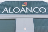 A large industrial building facade with the word 'ALOANCO' prominently displayed in white on a green background. Above the word, a multi-colored logo features a red, yellow, and orange triangular design. The building has green corrugated metal doors, and a small sign near the base indicates the presence of an alarm system.